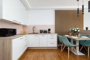 A modern kitchen with white cabinets, a black microwave, and a dining area with teal chairs and a wooden table. - Luxury Studio with Parking | Baltic Waves Resort | Near the Beach (Międzyzdroje)