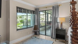 Ein Hotelzimmer mit großem Fenster, einem Sessel, einem Nachttisch mit Lampe und einem hölzernen Bettpfosten.