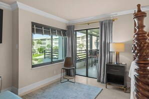 Ein Hotelzimmer mit großem Fenster, einem Sessel, einem Nachttisch mit Lampe und einem hölzernen Bettpfosten.