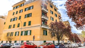 Un immeuble jaune à plusieurs étages, doté de balcons, avec des voitures garées et des arbres aux couleurs automnales.