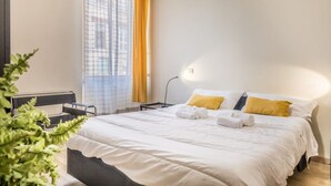 Une chambre d’hôtel avec un lit, un bureau, une chaise, une lampe et une fenêtre avec des rideaux.