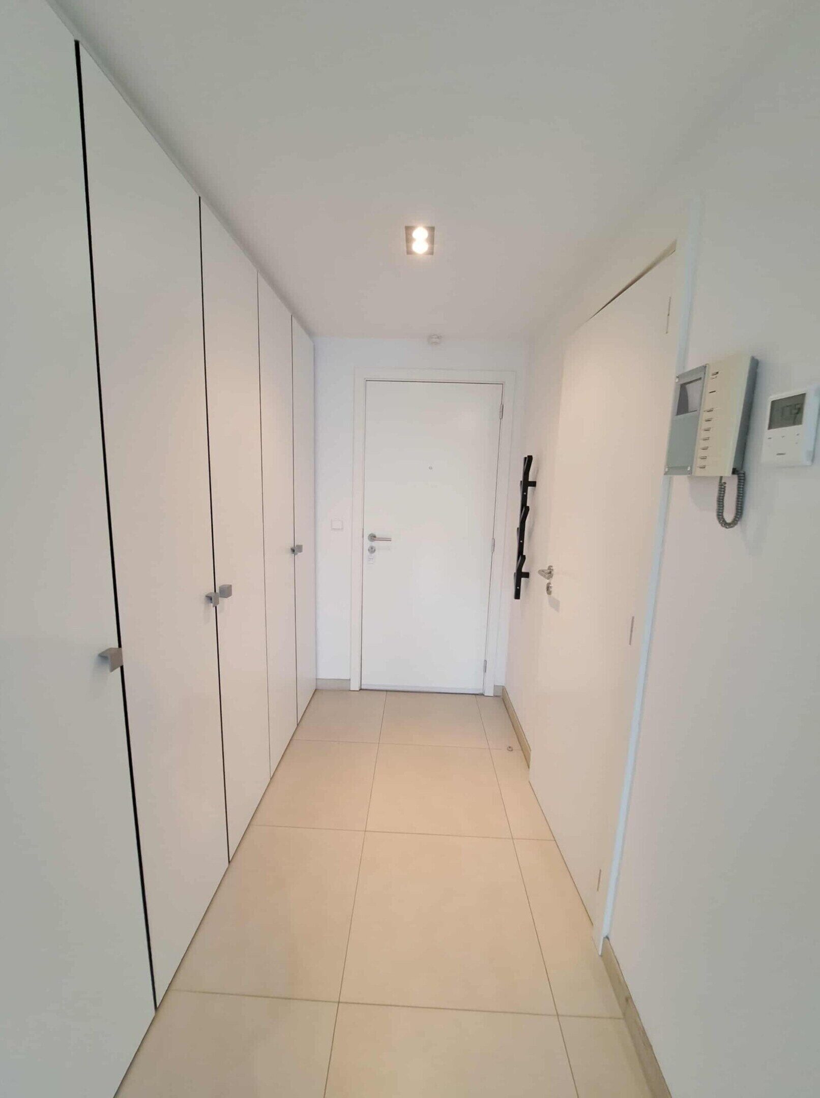 A hallway with white walls, a tiled floor, a door at the end, and a telephone on the wall.