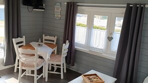 Quarto de canto com mesa de jantar, cadeiras, uma pequena mesa branca com bandeja, TV fixada na parede e janela com cortinas.