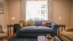 A living room with a grey sofa, two side tables with lamps, a patterned ottoman, and a window with curtains. - Challens Yarde is a large double fronted cottage, providing spacious, bright, and airy accommodation (Midhurst)