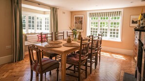A dining area with a wooden table, chairs, and a view of the outdoors through large windows. - Challens Yarde is a large double fronted cottage, providing spacious, bright, and airy accommodation (Midhurst)