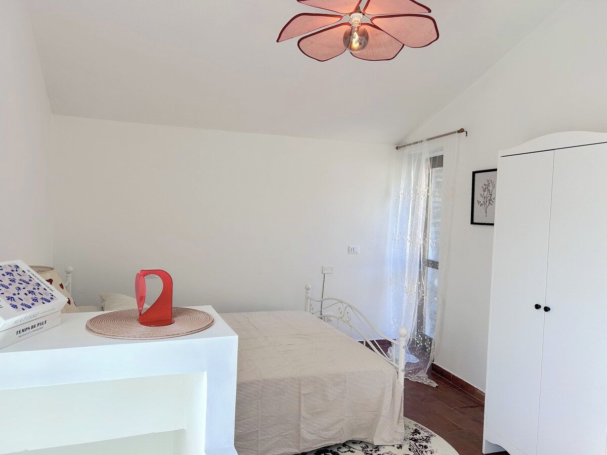 A bedroom with a bed, a white wardrobe, a red object on a table, and a ceiling fan.