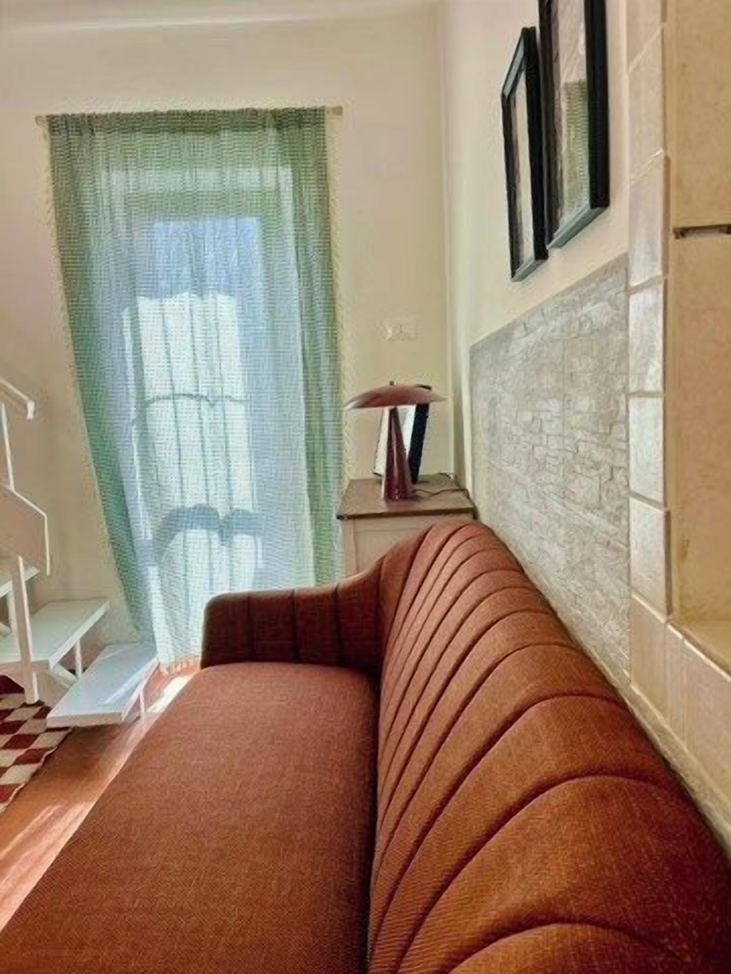 Ein Zimmer mit einem braunen Sofa, einem Fenster mit grünem Vorhang, einer Lampe auf einem Beistelltisch und einer gefliesten Wand.