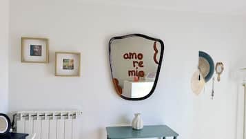 Een kamer met een radiator, een spiegel met de tekst 'amo', twee ingekaderde afbeeldingen, een klein tafeltje met een vaas, en een ventilator.