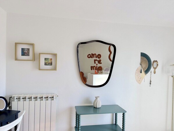A room with a radiator, a mirror with the word 'amo' on it, two framed pictures, a small table with a vase, and a fan.