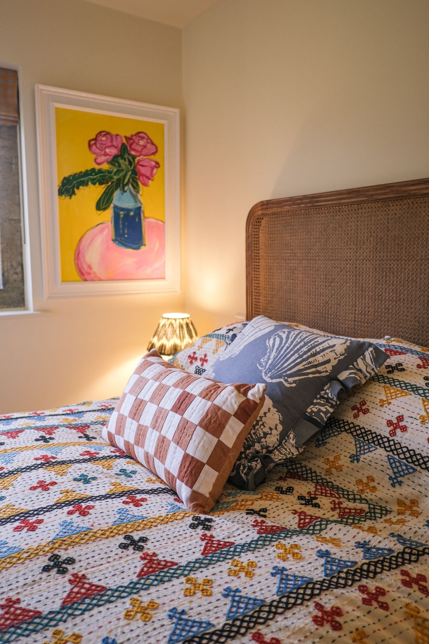 A bed with a colorful quilt, a checkered pillow, a decorative pillow, a lamp, and a painting on the wall.