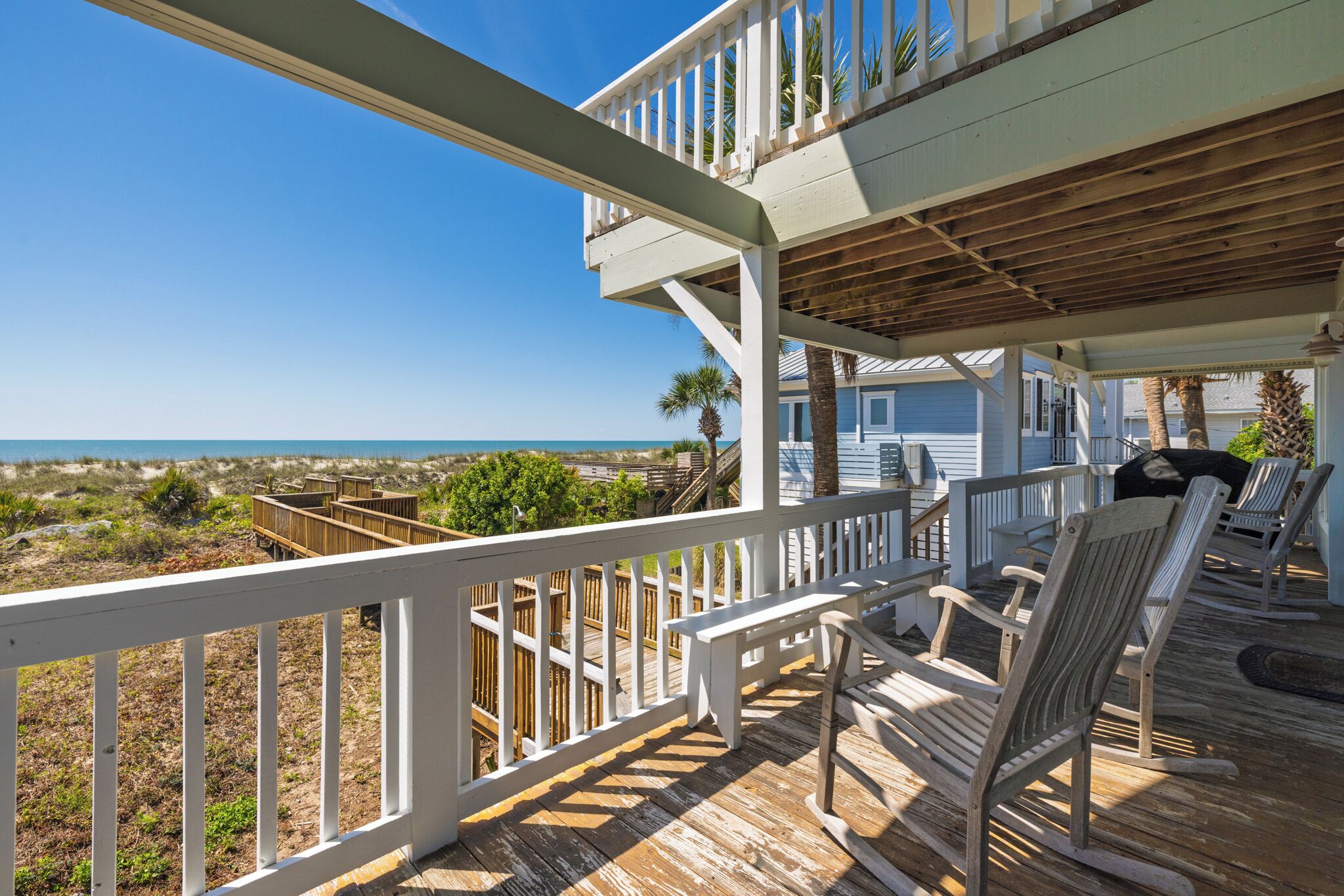 A wooden deck with white railings, a seating area with chairs, and a view of the ocean.