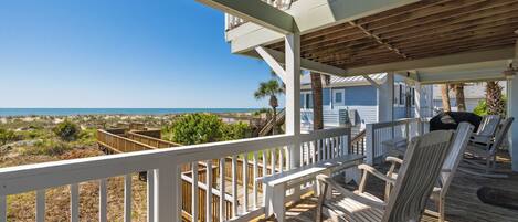 A wooden deck with white railings, a seating area with chairs, and a view of the ocean.