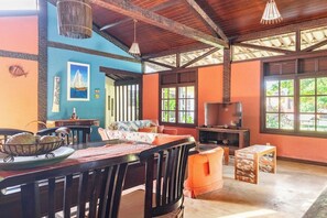 A spacious room with a high ceiling, a dining area with a table and chairs, a television, and large windows. - Ideal house for groups in Búzios | 2 houses + pool + staff + sports court + games room (Armação dos Búzios)