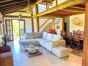 A spacious living area with a large sectional sofa, a glass coffee table, and a dining area with wooden chairs and a table. - Ideal house for groups in Búzios | 2 houses + pool + staff + sports court + games room (Armação dos Búzios)