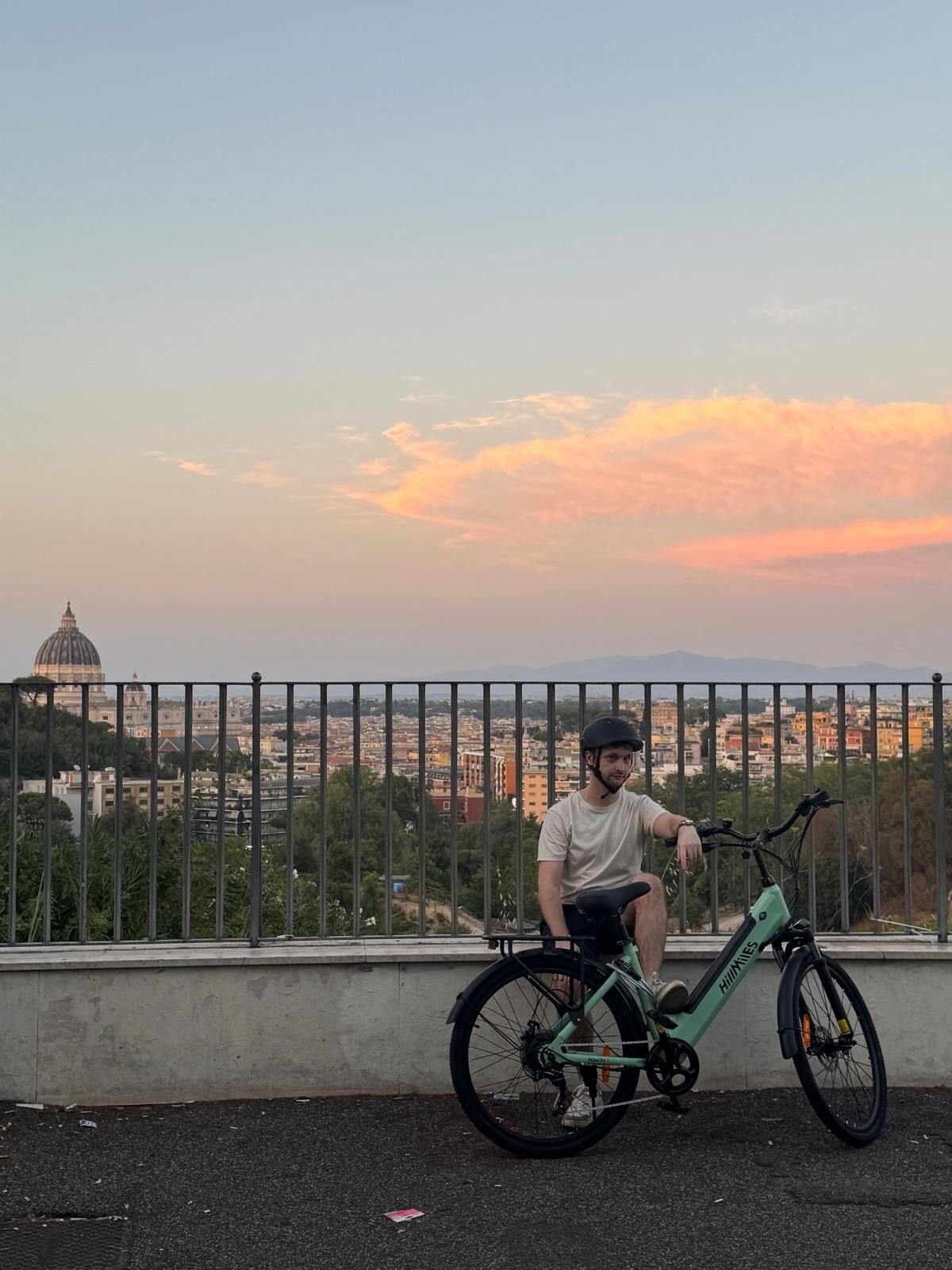 A person sitting on a green electric bike with a cityscape and a dome structure in the background.