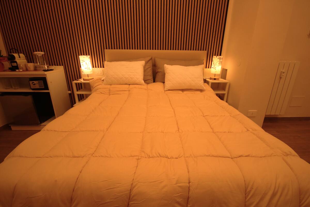Una cama con un edredón acolchado, lámparas de noche, una mesita y un escritorio con lámpara y cafetera.