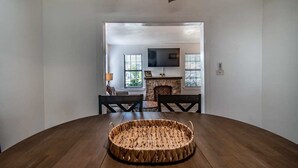 Un comedor con una mesa redonda de madera, sillas, una cesta tejida, una chimenea de piedra y un televisor montado en la pared.