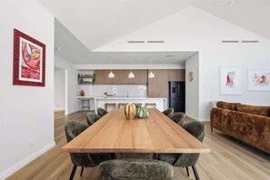 A modern dining area with a wooden table and chairs, a kitchen in the background, and a brown sofa. - Genoa House—Coastal Elegance at the Marina's Edge (Geographe)