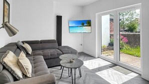 Un saló modern amb un sofà gris, una taula de centre rodona, un televisor penjat i una gran porta corredissa de vidre que dona a un espai exterior.