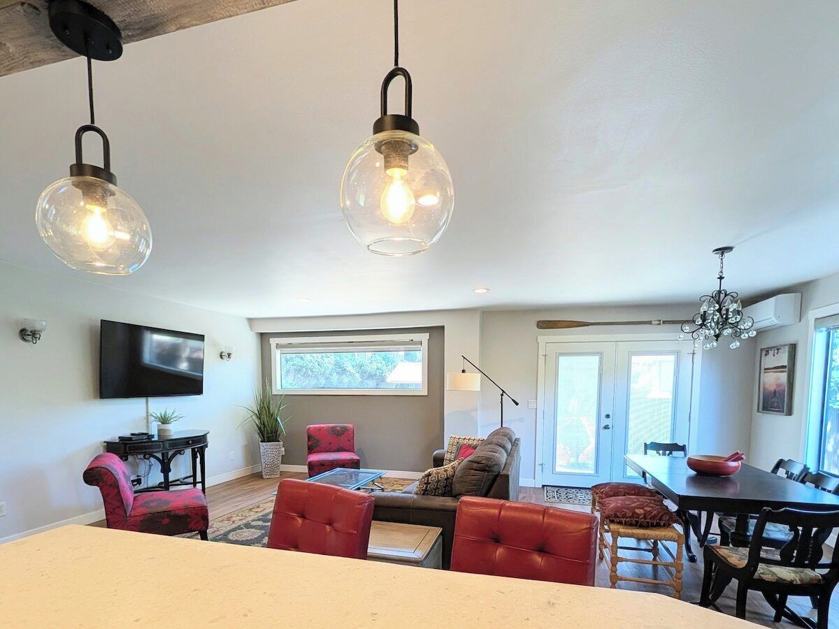 A modern living room with a dining area, a television mounted on the wall, and a large window providing natural light.