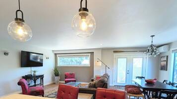 A modern living room with a dining area, a television mounted on the wall, and a large window providing natural light.