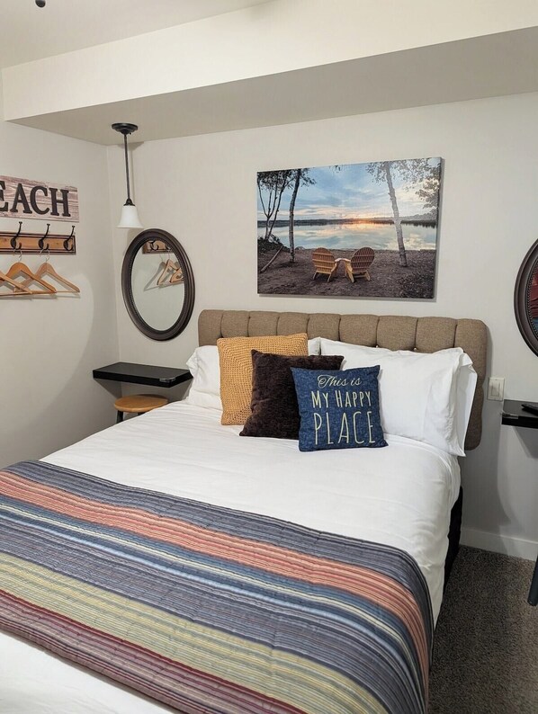 A neatly made bed with a striped bedspread, a pillow with the text 'This is my happy place,' and a painting of a beach scene above it.