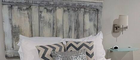 Una cama con ropa de cama blanca, almohadas estampadas, un cabecero rústico, una lámpara de noche y una pequeña mesita de cristal con una lámpara.