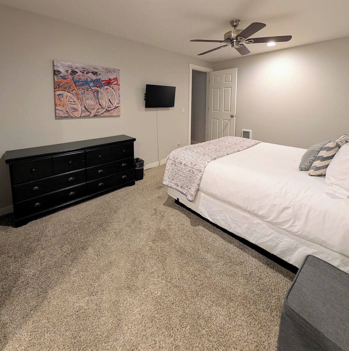 Une chambre à coucher avec un lit, une commode, un ventilateur de plafond, un téléviseur et une porte.