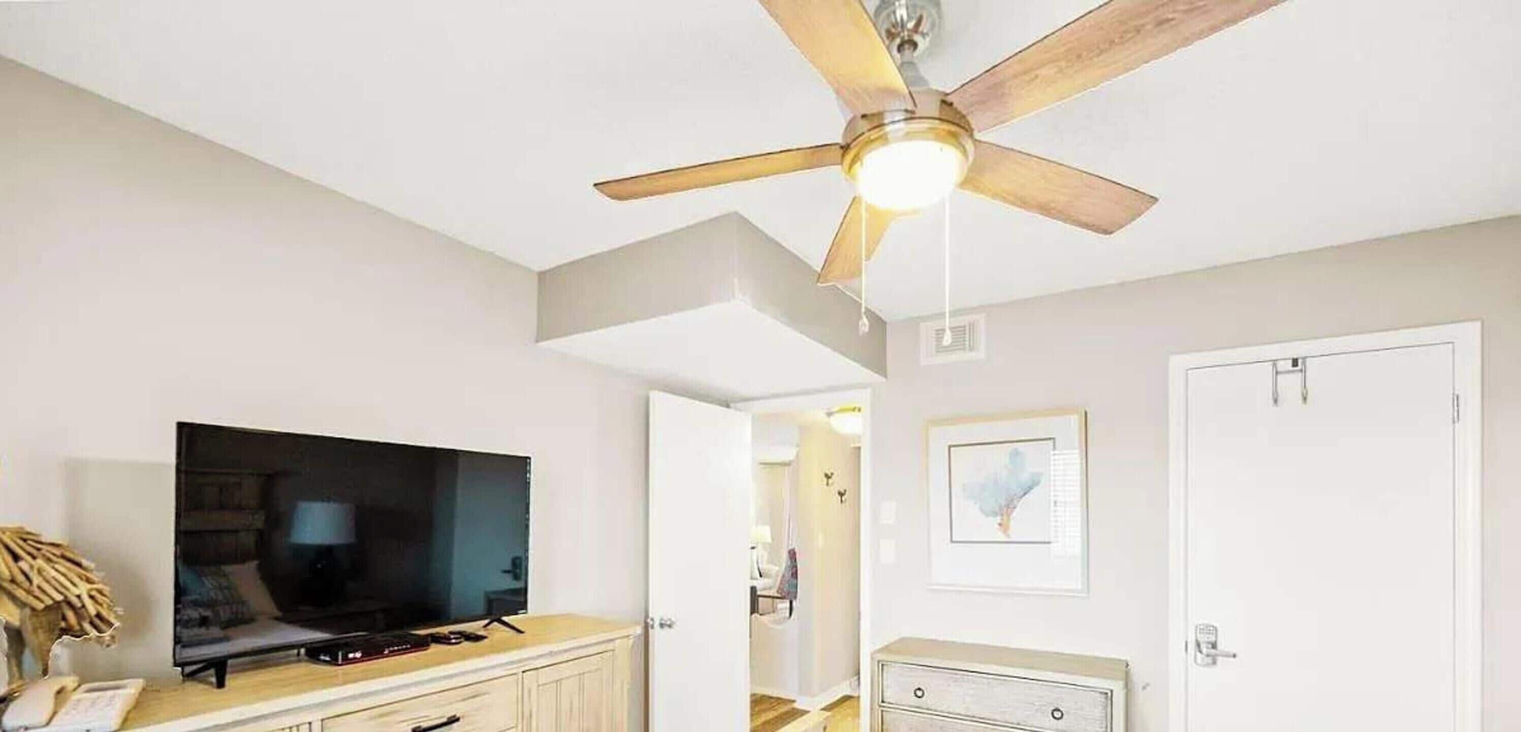 Una sala de estar con ventilador de techo, televisor de pantalla plana, mueble de madera, una puerta y un cuadro enmarcado en la pared.
