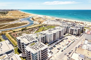 Ein moderner Wohnkomplex mit mehreren Gebäuden, einem Parkplatz und einem Strand im Hintergrund.