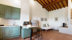 Una cocina con encimera blanca, armarios verdes, un taburete blanco, una cama blanca y un televisor sobre un soporte.