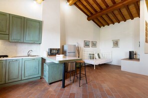 Una cocina con encimera blanca, armarios verdes, un taburete blanco, una cama blanca y un televisor sobre un soporte.