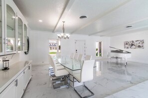 A spacious dining area with a glass-top table, white chairs, a piano, and a chandelier. - Sleeps 28 - Ocean View - Pearl on Ocean Dr, Combined Estate (Corpus Christi)