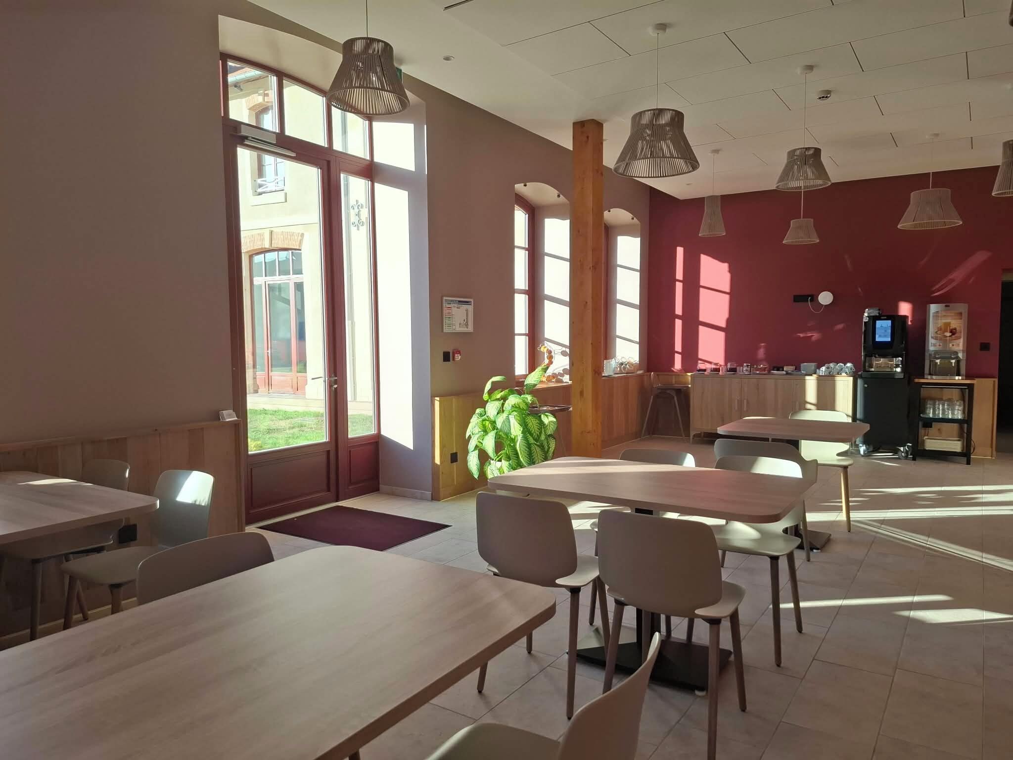 Un comedor espacioso con varias mesas y sillas, ventanas amplias que dejan entrar luz natural, y una zona de cocina al fondo.