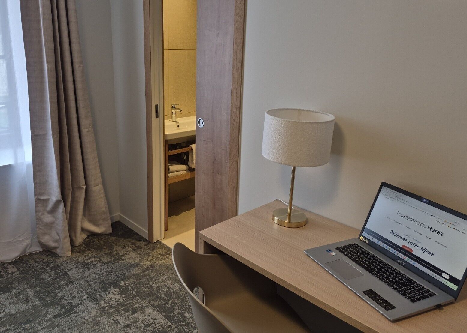 Habitación de hotel con escritorio, una computadora portátil, lámpara y baño visible a través de una puerta abierta.