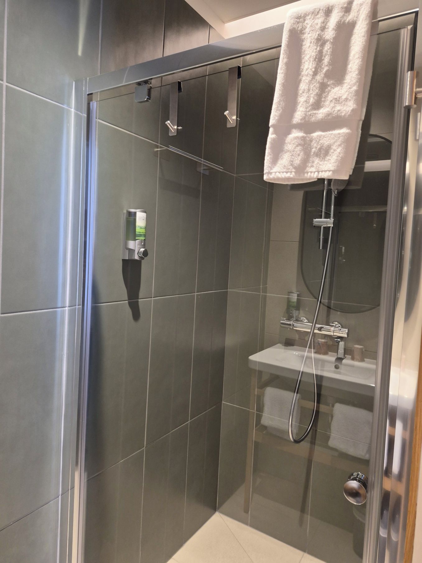 Un área de ducha moderna con mampara de vidrio, un cabezal de ducha manual, una toalla y un dispensador de jabón.