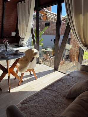 Ein gemütlicher Raum mit einem stuhlartigen Holzgestell, einem bequemen Sofa und einem Esstisch, dekoriert mit einem Blumenstrauß.
