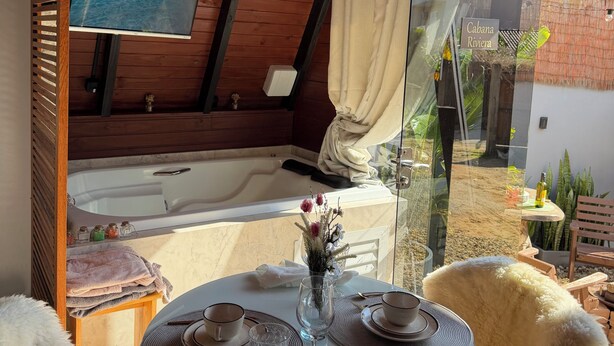 Ein gemütliches Zimmer mit Holzbalkendecke, einem großen Fenster mit Vorhängen, einer Badewanne, einem für zwei Personen gedeckten Tisch und Blick auf einen Garten.