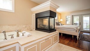 A bathroom with a large bathtub, a glass-enclosed fireplace, and a separate bedroom with a bed and a window. - Spacious 4-bedroom home with private beach,  dock, large yard, outdoor shower. (Kelowna)
