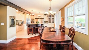A spacious dining area with a large wooden table and chairs, a kitchen with white cabinets, and a chandelier. - Spacious 4-bedroom home with private beach,  dock, large yard, outdoor shower. (Kelowna)
