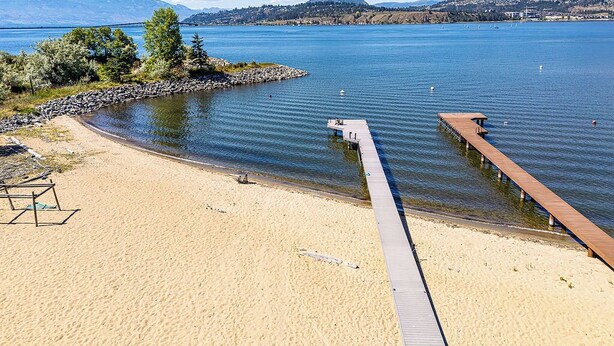 A beach with a wooden pier extending into calm water, surrounded by mountains and a clear sky. - Spacious 4-bedroom home with private beach,  dock, large yard, outdoor shower. (Kelowna)