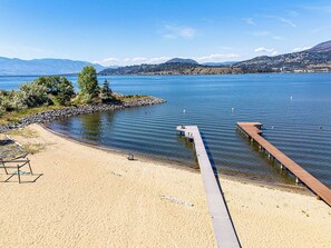 A beach with a wooden pier extending into calm water, surrounded by mountains and clear skies. - Spacious 4-bedroom home with private beach,  dock, large yard, outdoor shower. (Kelowna)