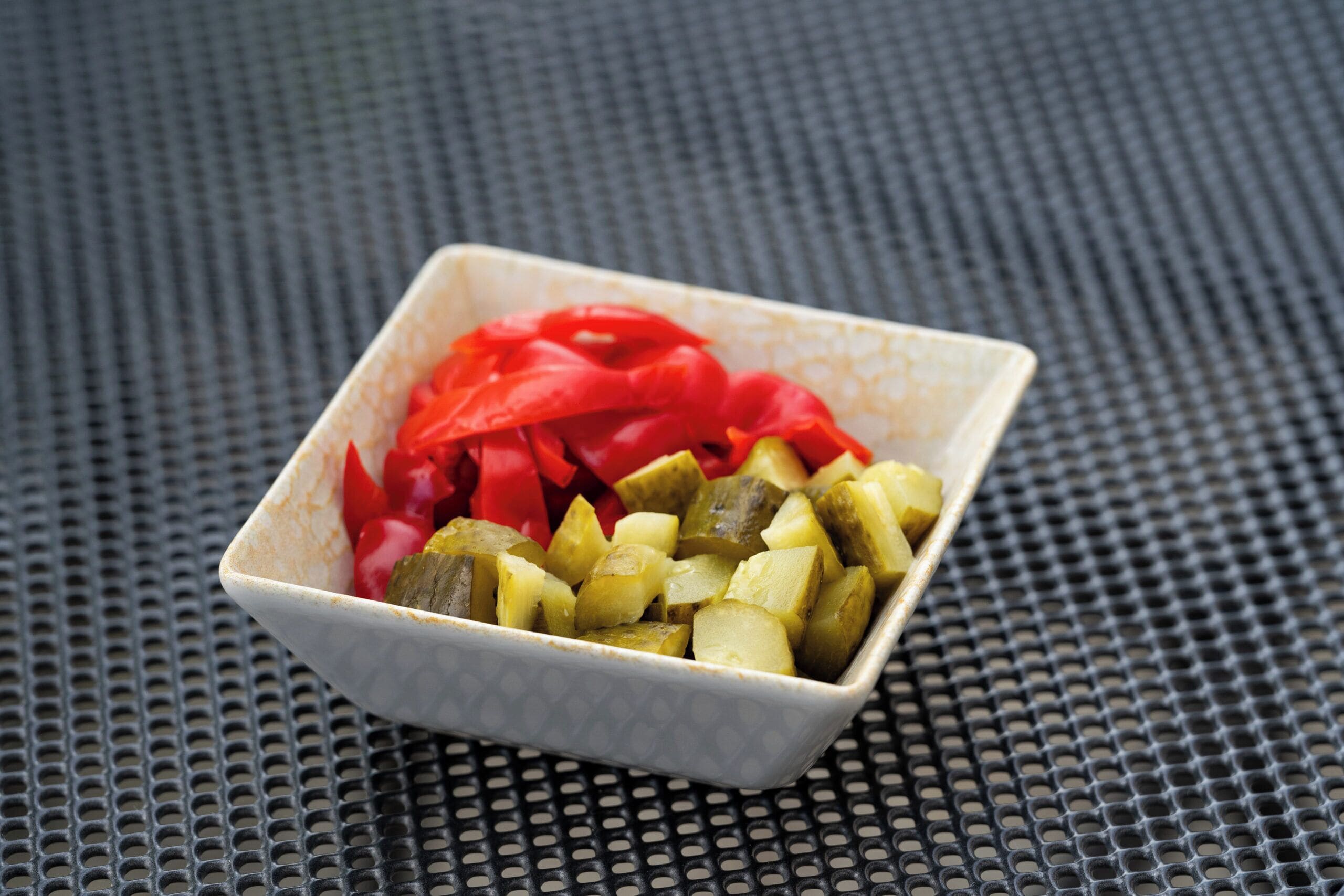A square ceramic bowl with diced pickles and red peppers on a textured surface.