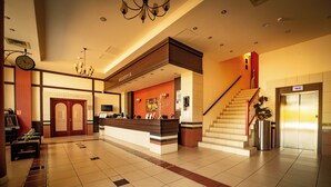 Zona de recepción de un hotel, con mostrador de recepción, un reloj, una escalera y un ascensor.