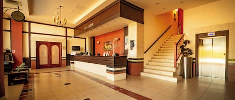 Zona de recepción de un hotel, con mostrador de recepción, un reloj, una escalera y un ascensor.