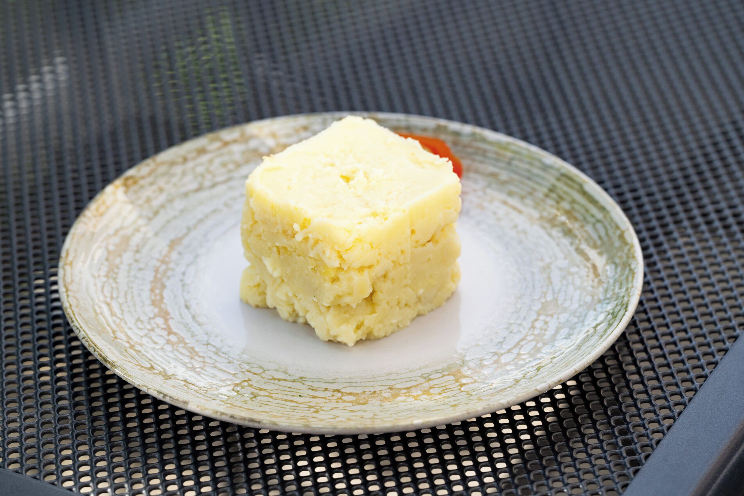 A square piece of food on a plate with a textured rim, placed on a perforated metal surface.