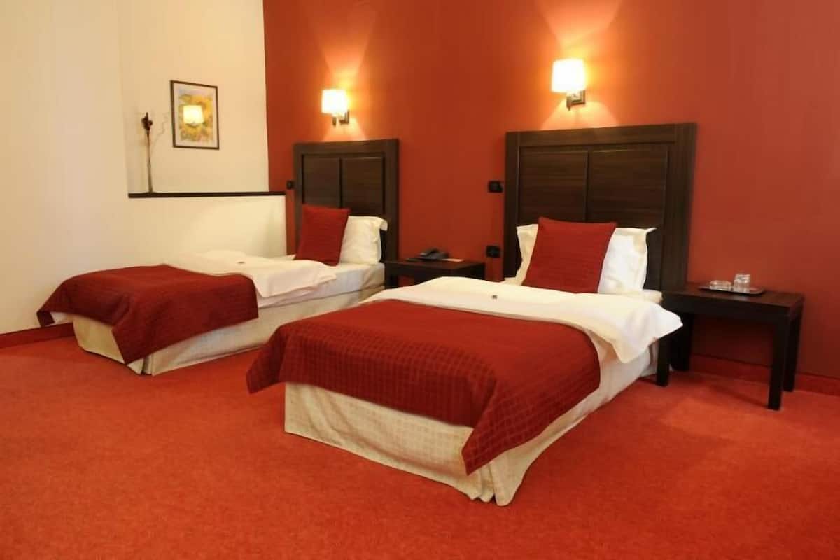 A hotel room with two beds, each with a red blanket, wooden headboards, and bedside tables with lamps.