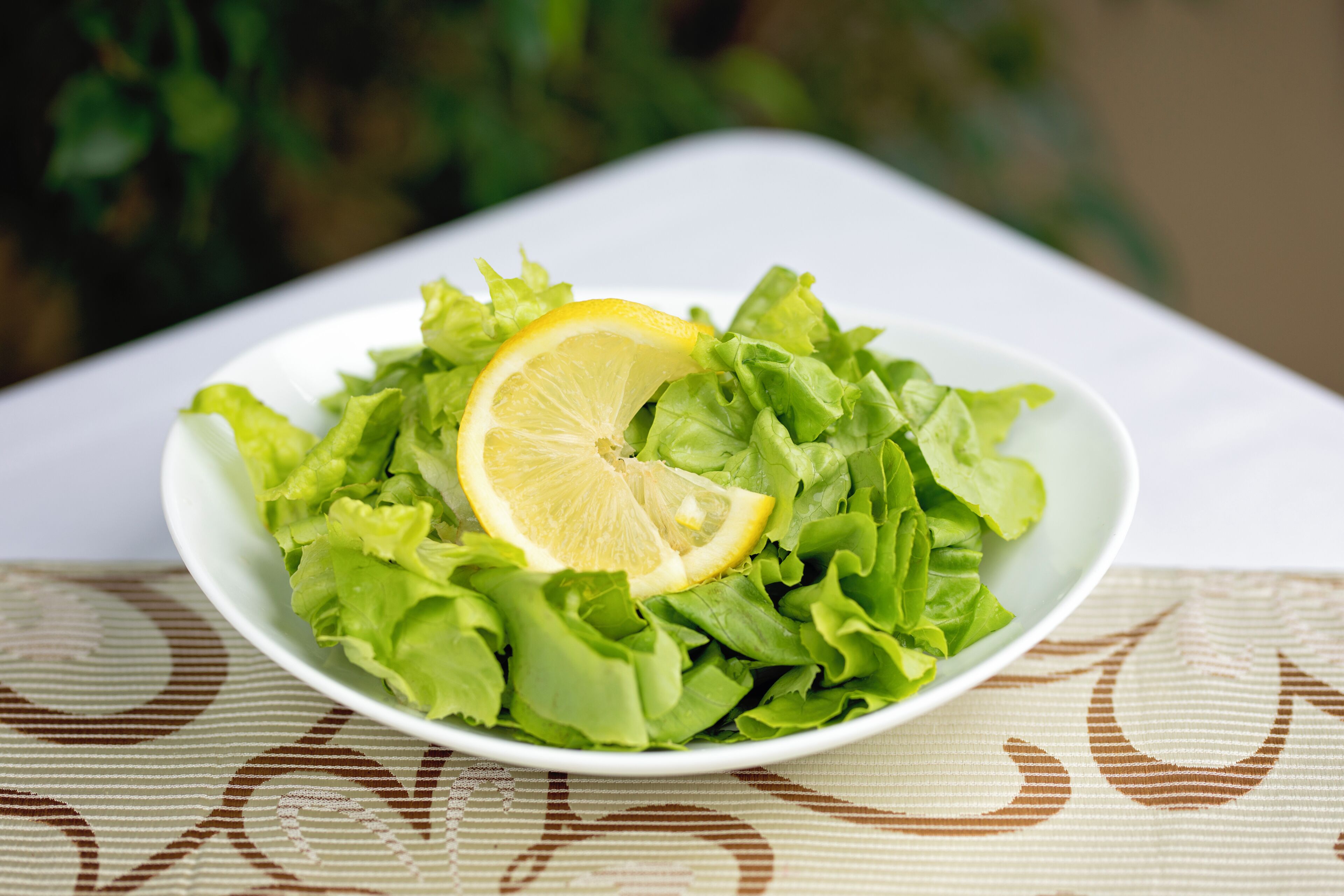 Un tazón blanco con ensalada verde y una rodaja de limón sobre un mantel estampado.