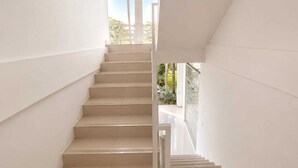 Un escalier aux murs blancs et à la rampe en verre, menant à un espace extérieur agrémenté de verdure.
