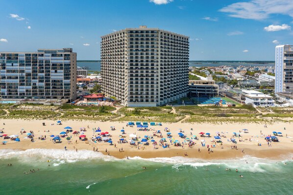 Ein Strand mit vielen Sonnenschirmen und Menschen, ein großes Hotelgebäude und im Hintergrund ein Gewässer.